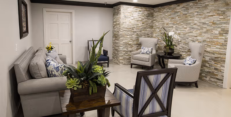 A cozy sitting area with a gray sofa adorned with patterned pillows, two beige armchairs with decorative cushions, and a wooden chair with striped upholstery. There is a wooden side table with a large green plant arrangement and a round black table with a white orchid and another plant. The walls feature a textured stone accent and a plain light-colored wall with a closed white door.