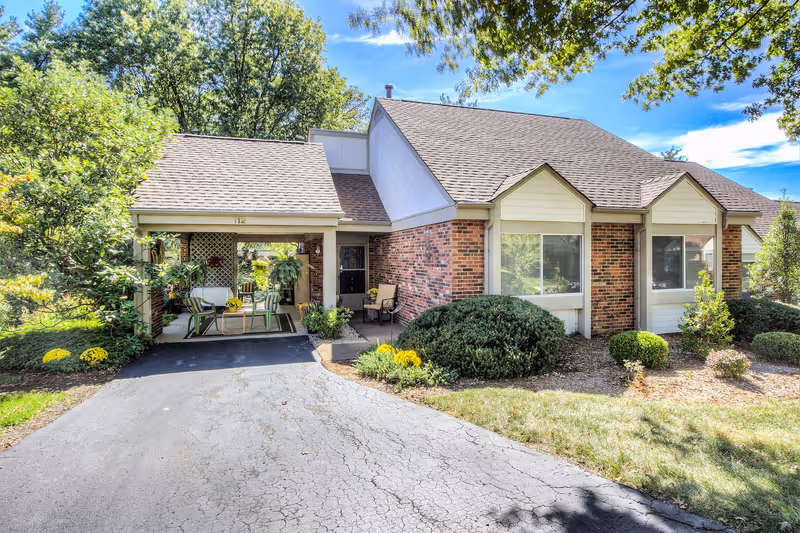 Exterior view of a single-story brick and siding building with a covered porch area furnished with chairs and plants. The driveway leads up to the porch, and there are well-maintained bushes and trees surrounding the building under a clear blue sky.