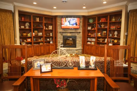 Cozy living room area with wooden bookshelves filled with books and decorative items, a stone fireplace with a mounted flat-screen TV above it, a patterned sofa, wooden chairs, and a wooden table displaying framed photos and decorative pieces.