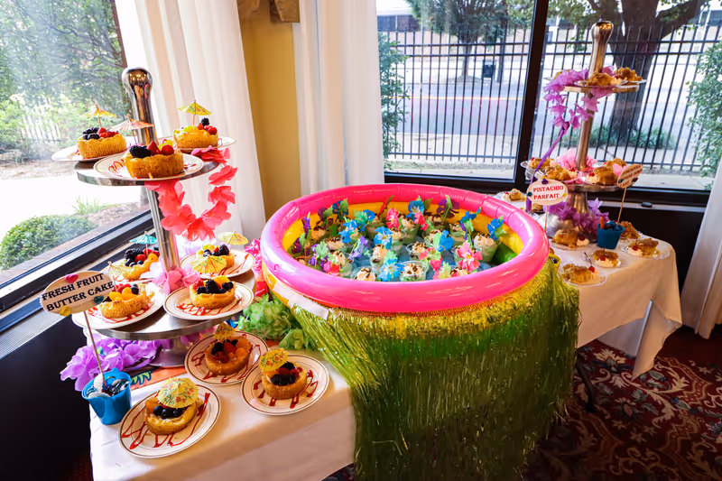 A dessert buffet table set up near windows with a view of trees and a street outside. The table features a tiered stand with fresh fruit butter cakes decorated with small umbrellas and berries, a small inflatable pool filled with colorful decorated parfait cups, and another tiered stand with pistachio parfaits. The table is decorated with green grass-like fringe and purple and pink leis.