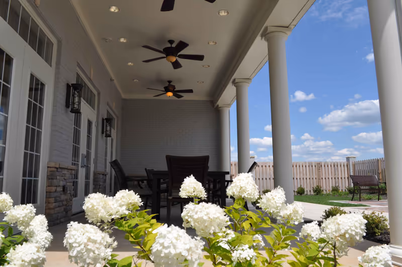 Covered outdoor patio area with white columns, ceiling fans, and a table with chairs. White flowers are in the foreground, and a wooden fence and blue sky with clouds are visible in the background.