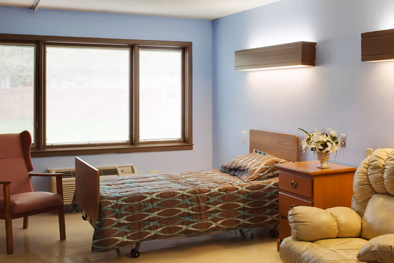 A single bed with a patterned brown and blue bedspread in a room with light blue walls. Next to the bed is a wooden nightstand with a vase of white flowers. There is a beige recliner chair in the foreground and a red armchair near a large window with blinds. The room has a simple, clean, and cozy appearance.
