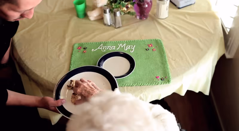 A birthday cake labeled Anna May on a table with people gathered.
