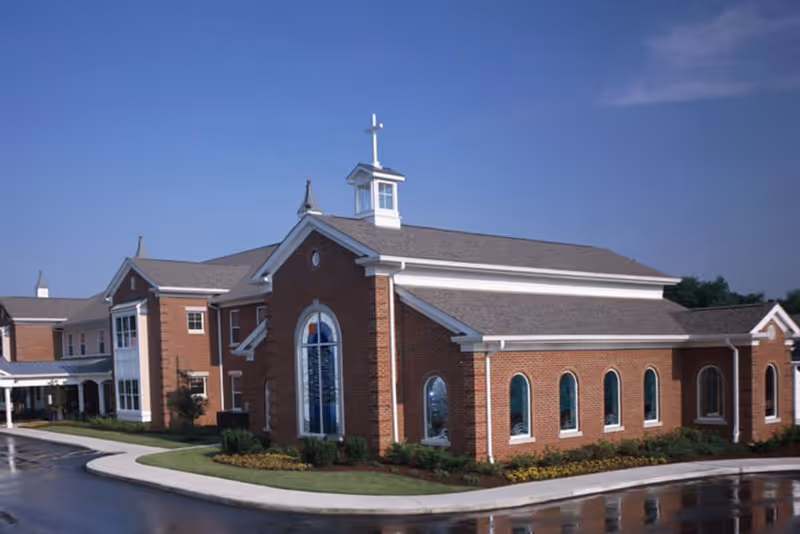 Brick senior living facility building with a chapel-like section featuring arched stained-glass windows and a small cross on the roof under a blue sky.