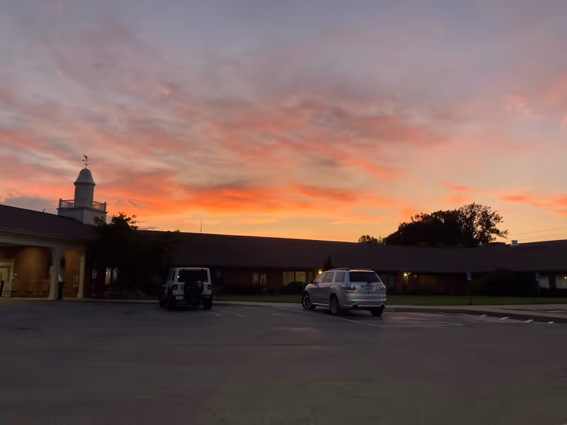 Sunset skies over a single-story nursing facility exterior with a parking lot in the foreground.