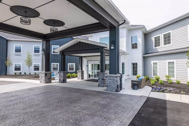 Entrance area of Provision Living at Crown Ridge featuring a covered drop-off zone with stone pillars and overhead lighting. The building exterior is clad in light gray and dark blue siding with multiple windows. There are small landscaped areas with shrubs and flowers near the entrance, and outdoor seating including a bench and a chair with cushions.