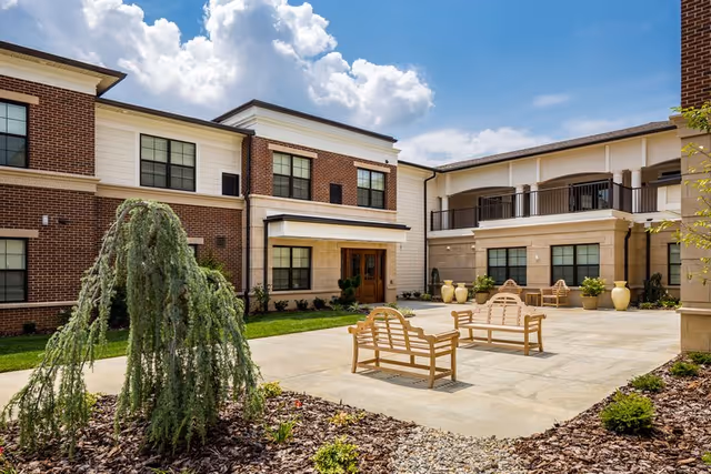 Outdoor courtyard area of a senior living facility with wooden benches, potted plants, and a small tree in a landscaped bed. The building has a brick and beige exterior with multiple windows and a covered entrance. The sky is partly cloudy.