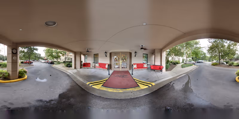Covered entrance area of The Lafayette senior living facility with a maroon welcome mat, red benches on either side, ceiling fans, and glass double doors. The driveway curves around the entrance with parked cars and trees visible in the background.