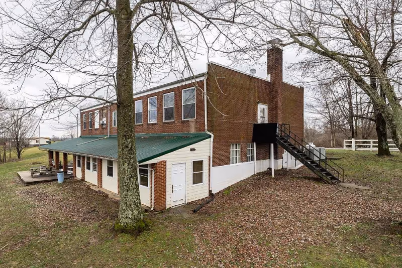 Exterior view of a two-story brick building with a green metal roof over a lower level extension. There are several windows on both floors, a black metal staircase leading to a door on the upper floor, and leafless trees surrounding the building. The ground is covered with fallen leaves and grass.