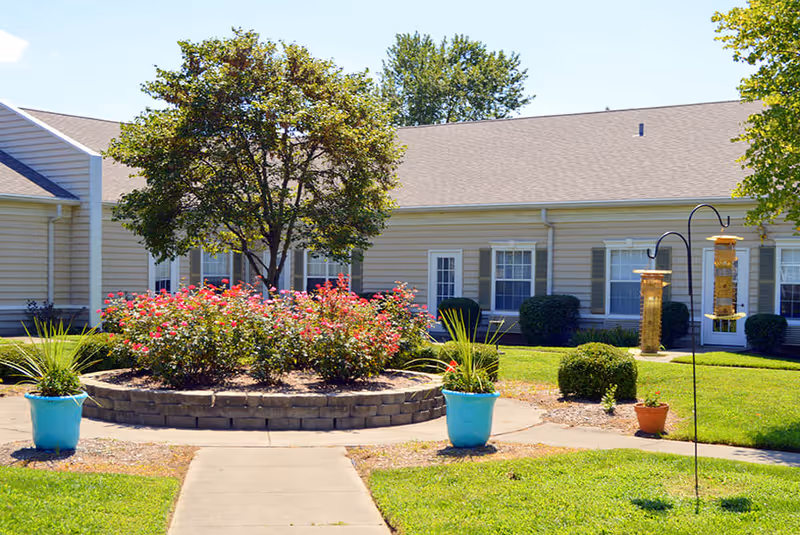 Outdoor garden area at Morning Pointe of Owensboro featuring a circular raised flower bed with blooming red flowers and a tree in the center, surrounded by a paved walkway. There are two blue planters with green plants on either side of the walkway and bird feeders hanging on metal hooks. The background shows a beige building with white-framed windows and green shutters.