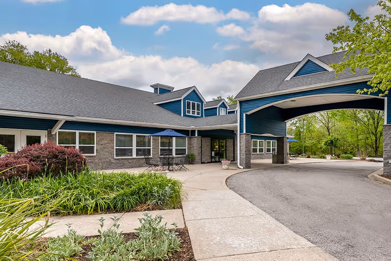 Exterior view of Brookdale Stonestreet facility showing a covered driveway entrance, blue siding with stone accents, windows, outdoor seating with tables and umbrellas, and landscaped greenery under a partly cloudy sky.