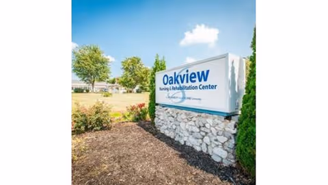 Outdoor view of a sign for Oakview Nursing and Rehabilitation Center set on a stone base with bushes and trees in the background under a blue sky with a few clouds.