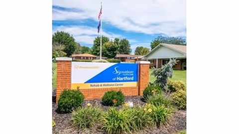 Outdoor view of the entrance sign for Signature HealthCARE of Hartford Rehab & Wellness Center, surrounded by landscaping with bushes and flowers, with a building and flagpole in the background under a partly cloudy sky.