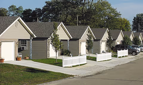 Row of beige townhouse-style retirement cottages with white fences along a street.