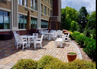 Outdoor paved patio beside a multi-story assisted living building with white tables and chairs and surrounding landscaping.