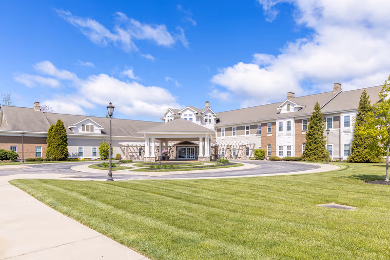 Exterior view of a senior living community building with a circular driveway, well-maintained green lawn, trees, and a partly cloudy blue sky.