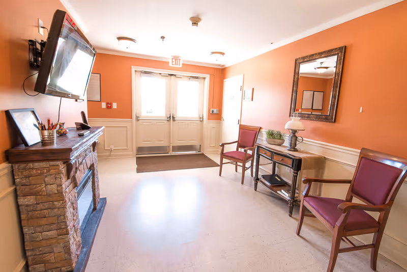 A bright hallway with orange walls and white trim, featuring a stone fireplace with a mounted TV above it on the left side. Two wooden chairs with red cushions are placed on the right side next to a wooden console table with a lamp, a plant, and some books. Double doors with windows and an exit sign above them are at the end of the hallway.