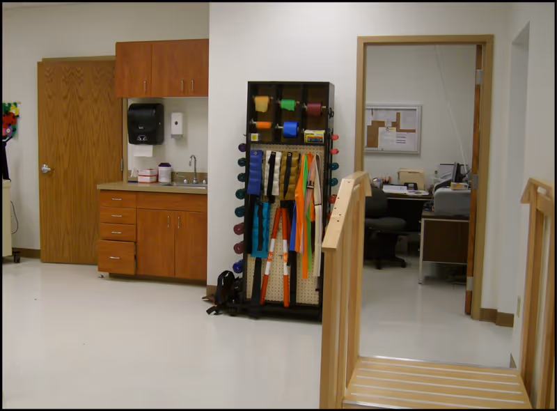 Interior room with a wooden door, a countertop with cabinets, a sink, and a wall-mounted soap dispenser. Next to the countertop is a rack holding various colored resistance bands and exercise equipment. To the right, there is an open doorway leading to an office with a desk, chair, computer, and bulletin board. In the foreground, there is a wooden ramp with handrails.