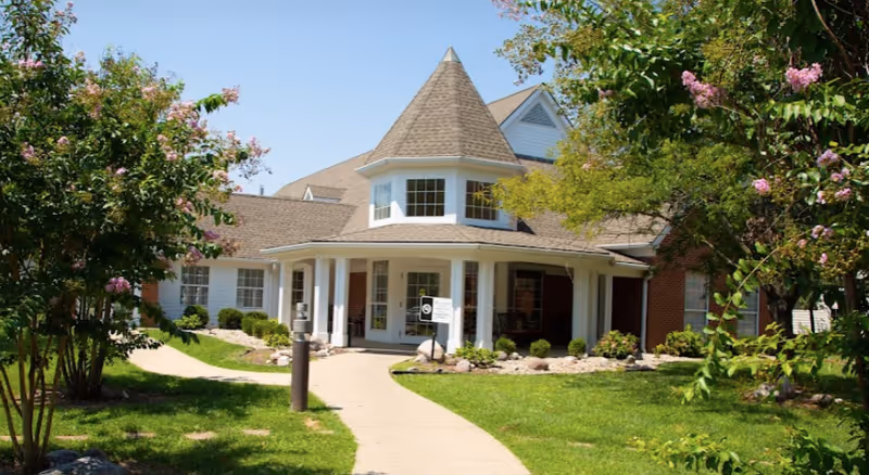 Front entrance of a red-brick senior living building with a conical turret, covered porch, walkway and landscaped lawn with flowering trees.