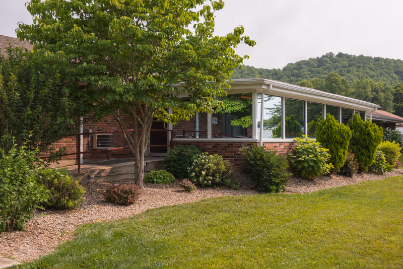 Exterior view of a brick building with large windows, surrounded by green shrubs, trees, and a well-maintained lawn with a backdrop of forested hills under a cloudy sky.