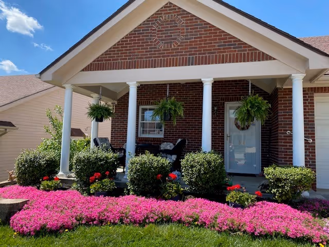 Front porch of a brick house with white columns, hanging ferns, seating and a bright pink flowerbed in front.