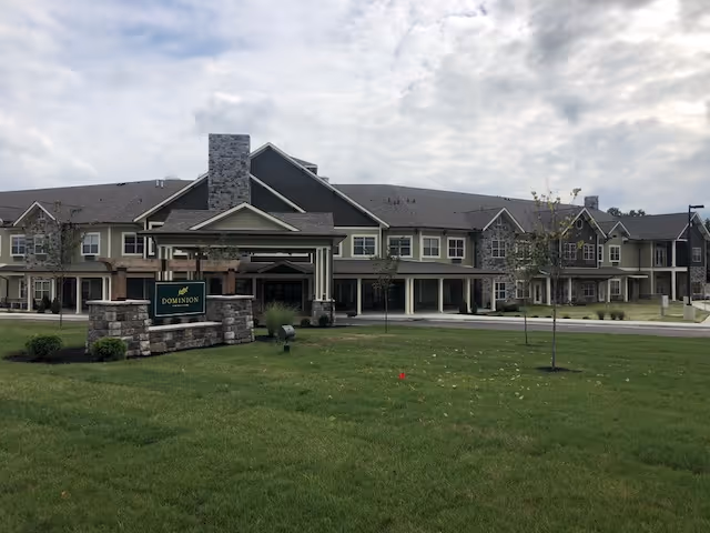 Exterior view of Dominion Senior Living of Frankfort, a large two-story building with a stone and siding facade, a covered entrance, and a well-maintained lawn in the foreground under a cloudy sky.