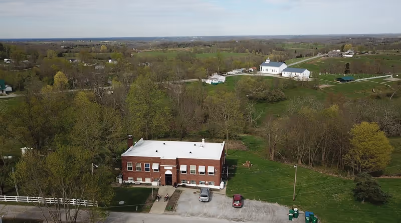 Aerial view of a red brick building with a white roof, surrounded by green trees and grass. There are a few cars parked in front of the building and a white fence on the left side. In the background, there are open fields, scattered houses, and a white church with a steeple.