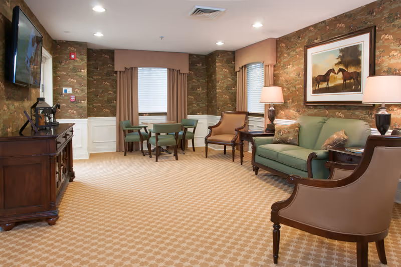 Senior living facility lounge with green sofa, armchairs, tables, lamps, and framed horse painting.